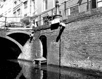 Gezicht op gedeelten van de voorgevels van de huizen Drift 23-25 te Utrecht, vanaf het water van de gracht, uit het westen met op de voorgrond de gracht met kademuur en keldertoegang; links de aanzet van een brug naar het huis Drift 6 in 1976. Bron: Het Utrechts Archief, catalogusnummer: 56355.
