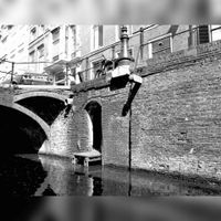 Gezicht op gedeelten van de voorgevels van de huizen Drift 23-25 te Utrecht, vanaf het water van de gracht, uit het westen met op de voorgrond de gracht met kademuur en keldertoegang; links de aanzet van een brug naar het huis Drift 6 in 1976. Bron: Het Utrechts Archief, catalogusnummer: 56355.