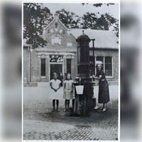 Twee meisjes en een mevrouw poserend bij de pomp op het Plein, op de achtergrond de voorgevel van de openbare lagere school ''t Groentje in 1930. Bron: Regionaal Archief, 353, 40305, 47.